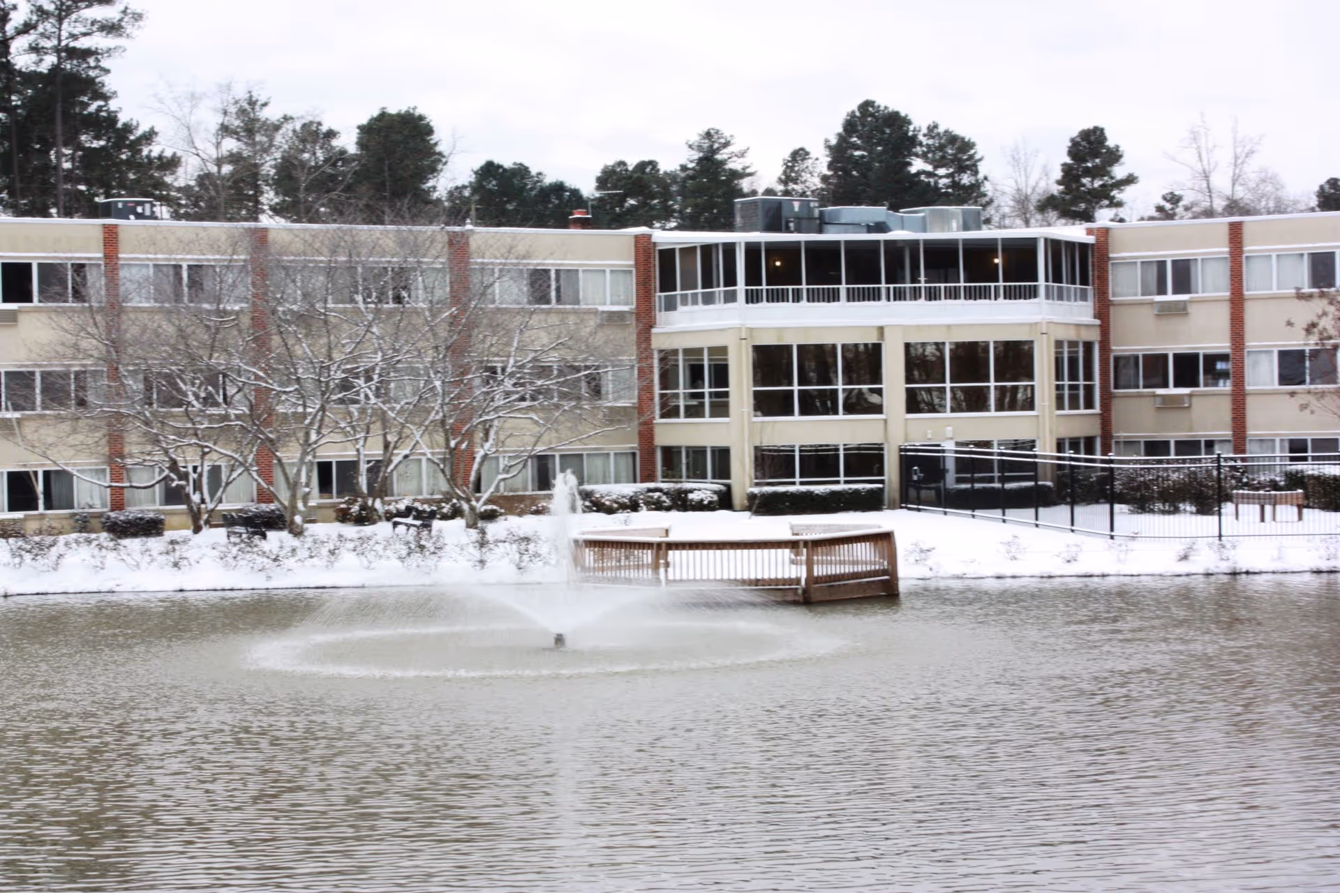 Snow-covered multi-story senior living building seen across a pond with a central fountain and a small wooden dock.