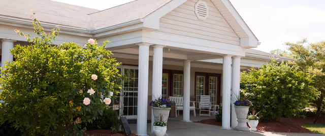 Front entrance of Gables Care Center featuring a covered porch with white columns, two white rocking chairs, potted plants with flowers, and surrounding green bushes and trees under a partly cloudy sky.