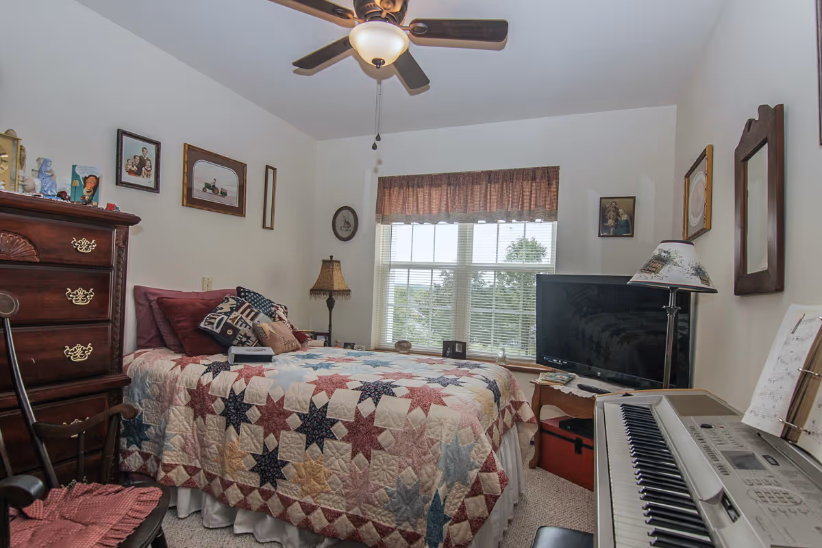 A cozy bedroom with a bed covered in a colorful star-patterned quilt, several pillows, a wooden dresser with decorative items, a ceiling fan, a window with blinds and a valance, a TV on a small table, a floor lamp, framed pictures on the walls, and an electronic keyboard with sheet music.
