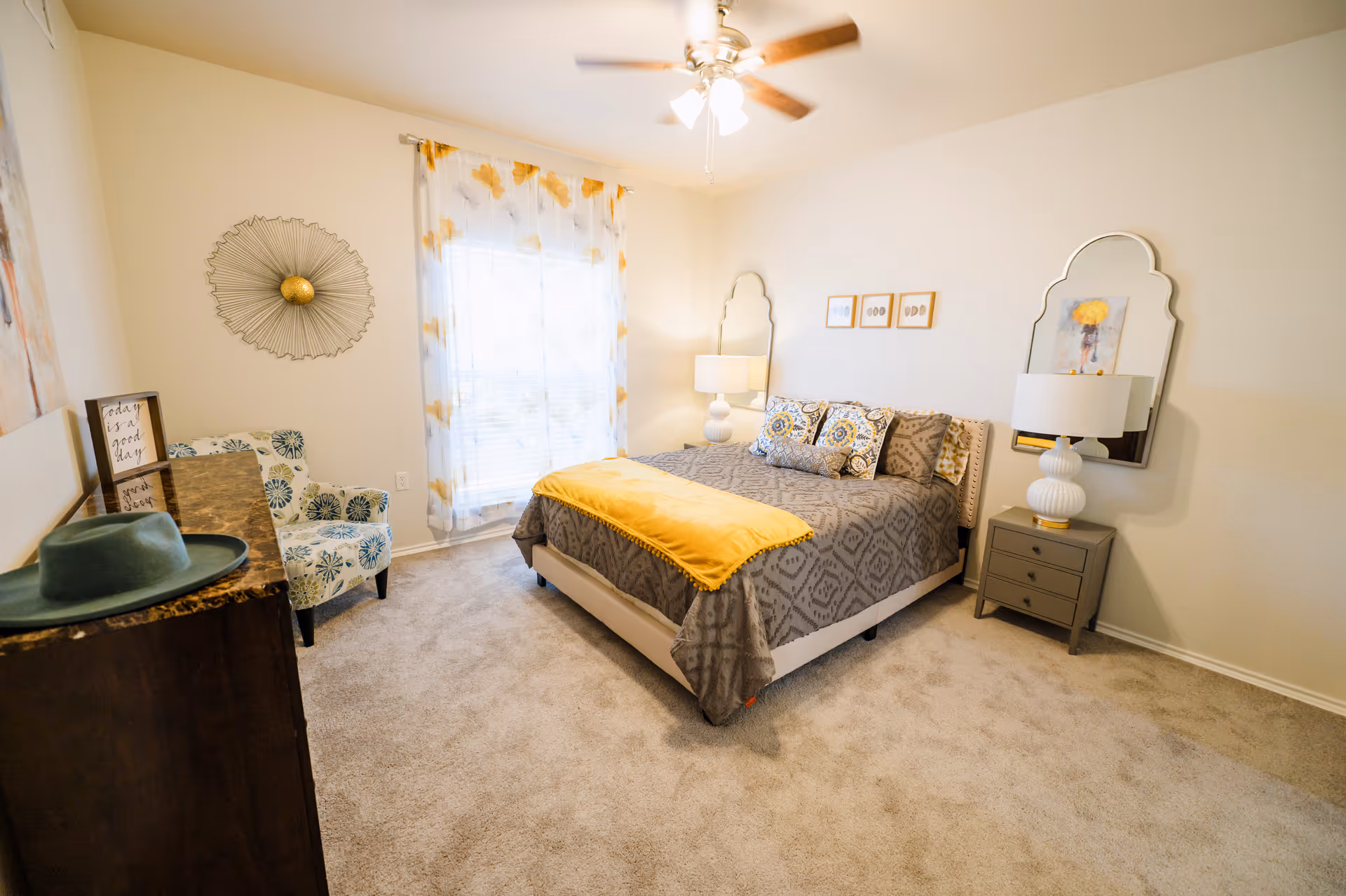 Bright furnished bedroom with a bed, nightstands and lamps, a patterned armchair, dresser, and a window with sheer curtains.