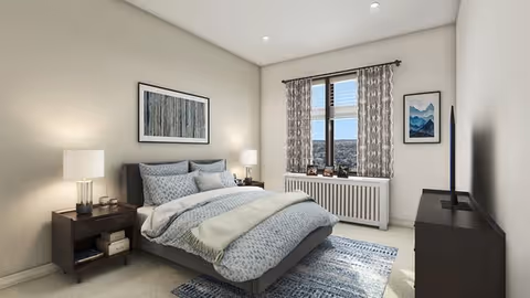 A cozy bedroom with a neatly made bed featuring blue and white bedding, two bedside tables with lamps, a window with patterned curtains, a radiator below the window, a framed picture on the wall above the bed, another framed picture on the adjacent wall, and a dark-colored dresser with a TV on top.