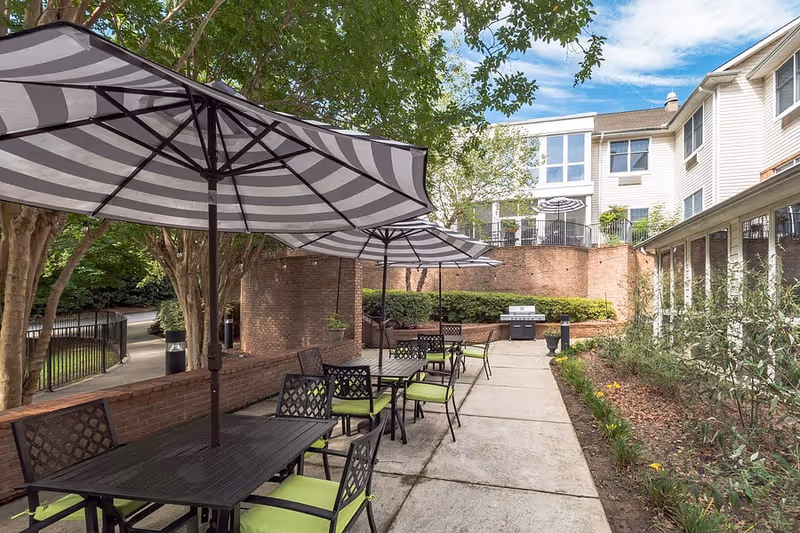 Outdoor patio area with several black metal tables and chairs with green cushions, shaded by large striped umbrellas. The patio is surrounded by brick walls and greenery, with a building in the background under a partly cloudy sky.