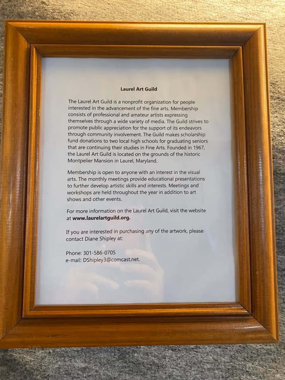 A framed printed notice about the Laurel Art Guild in a wooden frame mounted on a wall.
