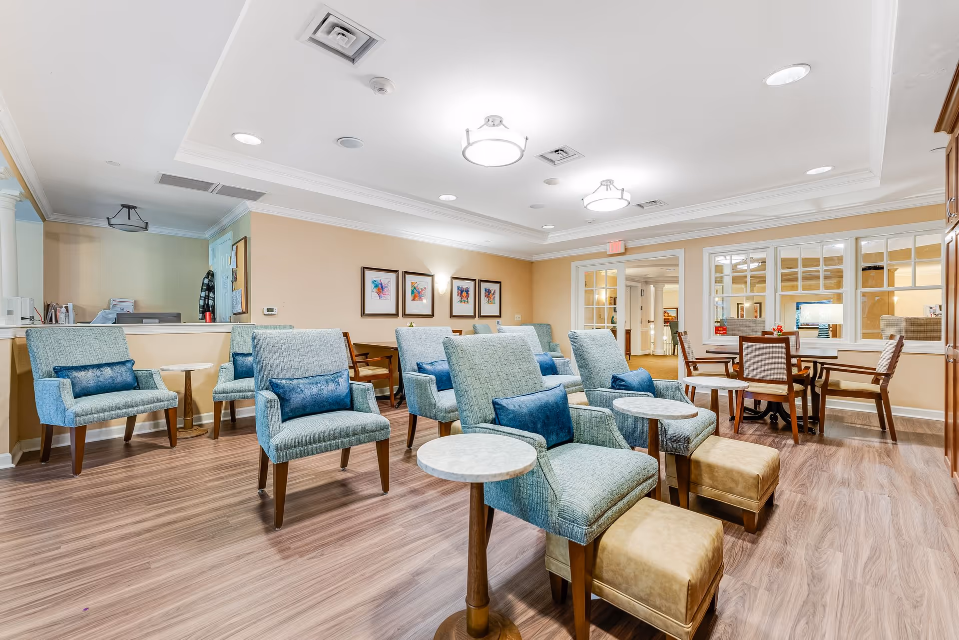 A bright and spacious common area in a senior living facility with multiple light blue upholstered armchairs, each paired with a small round side table and a cushioned footrest. The room has wood flooring, beige walls adorned with framed artwork, and ceiling lights. In the background, there is a table with chairs near a windowed wall and a reception desk area to the left.