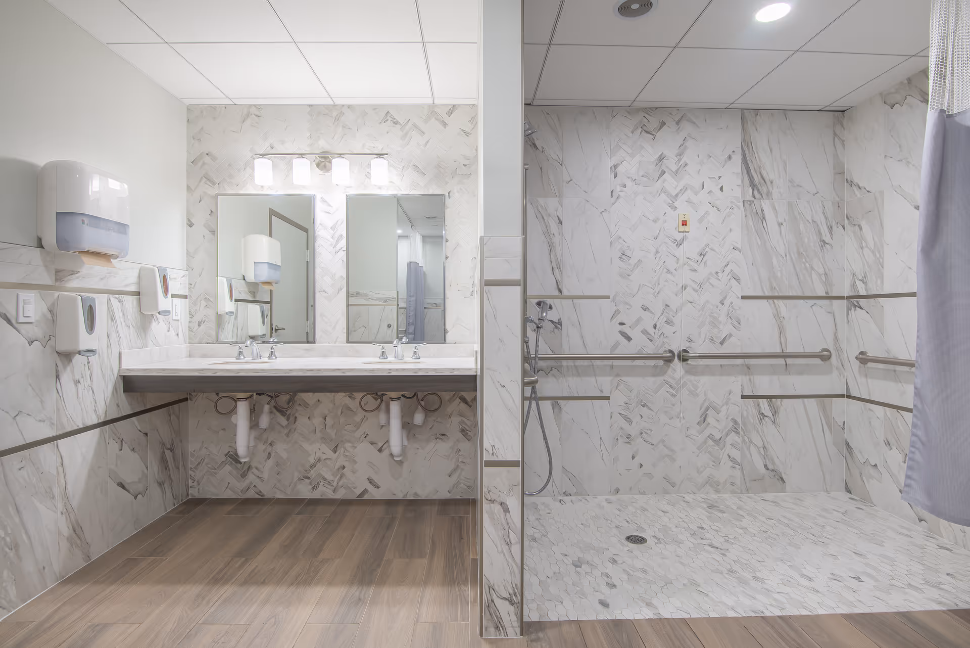 A clean, modern bathroom with a large vanity featuring two sinks and mirrors above. The walls and shower area are decorated with light-colored marble tiles. The shower is equipped with multiple grab bars and a handheld showerhead, and there is a shower curtain partially visible on the right. The floor has wood-like tiles, and there are soap and paper towel dispensers mounted on the wall.