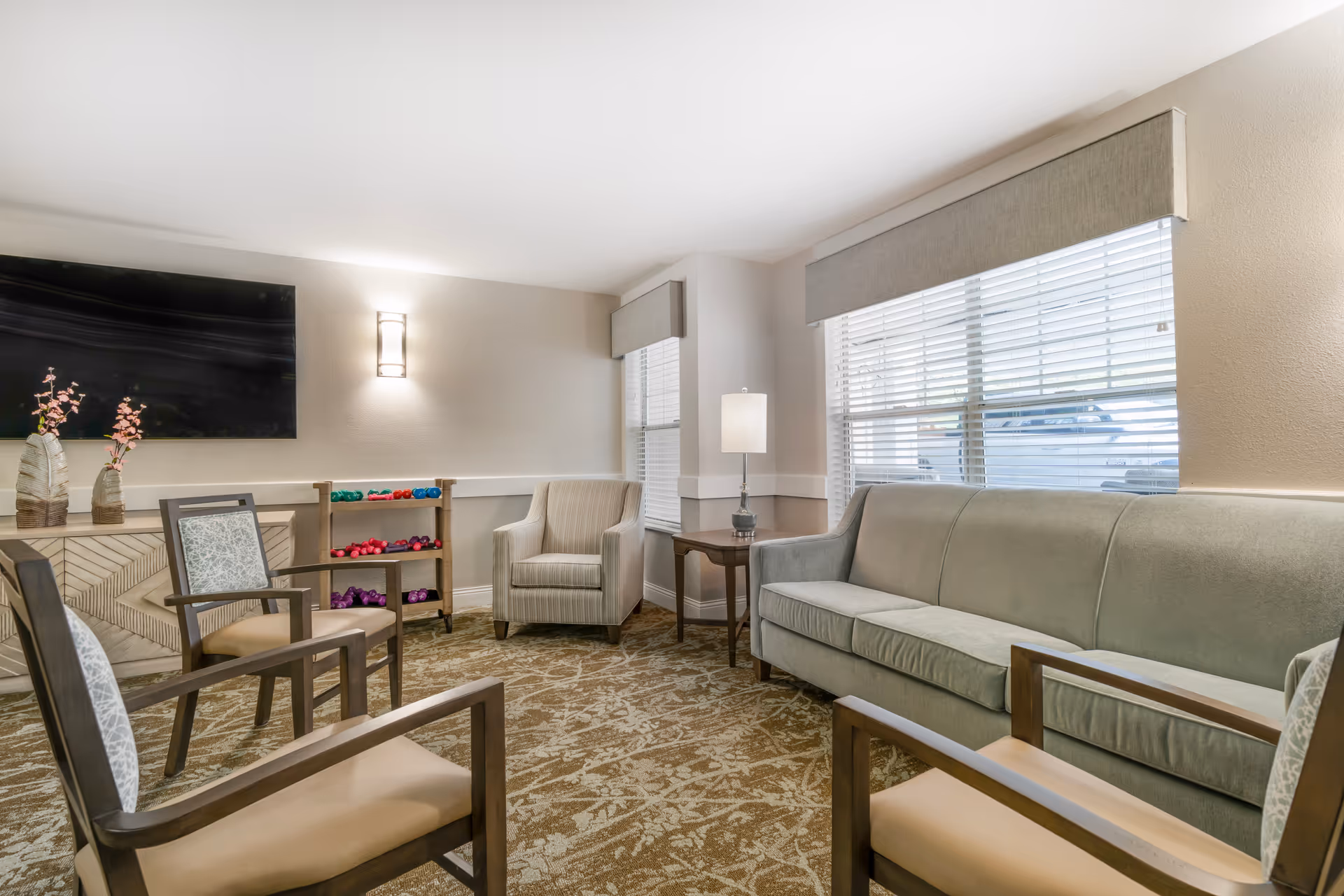 A well-lit living room with a large window covered by blinds, a green sofa, four wooden chairs with cushions, a small wooden side table with a lamp, a wall-mounted flat screen TV, a cabinet with decorative vases, and a rack holding colorful dumbbells.