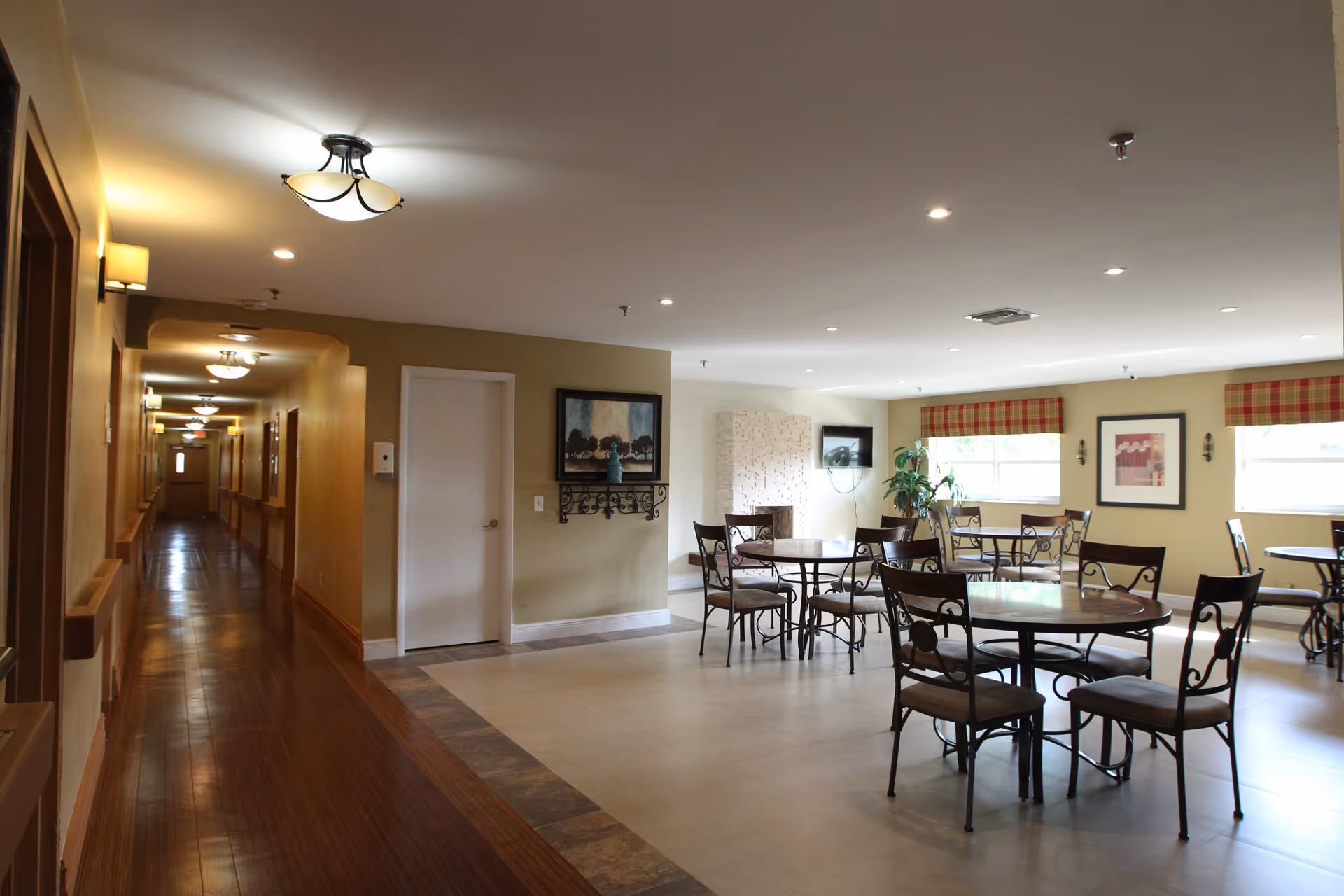 Interior view of a senior living facility showing a long hallway with wooden flooring and multiple doors on the left. To the right, there is a common area with several round tables and chairs, a wall-mounted TV, a fireplace, and windows with plaid valances allowing natural light to enter.