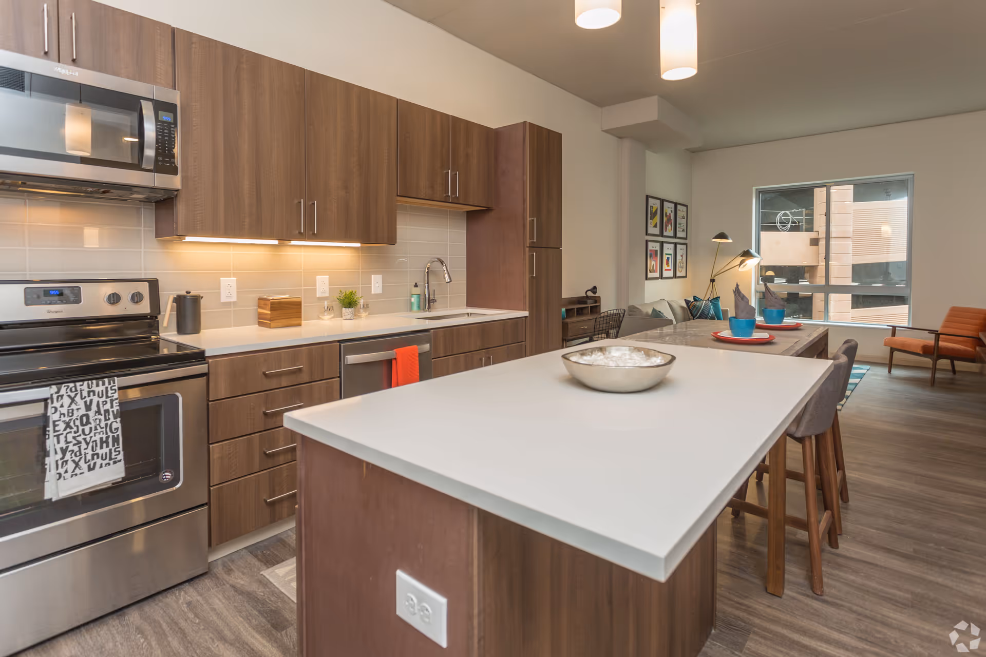 Modern kitchen with wooden cabinets, stainless steel appliances including a microwave and stove, a white countertop island with a bowl, and a dining area with table settings and chairs near a large window.