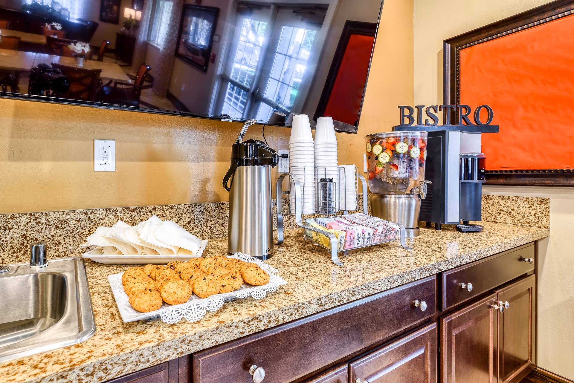 Countertop with a plate of cookies, napkins, a coffee dispenser, stacked disposable cups, a container with assorted sweeteners, and a beverage dispenser filled with water and slices of cucumber and strawberries. A large TV is mounted on the wall above the counter, and a sign that reads 'BISTRO' is displayed next to the beverage dispenser.