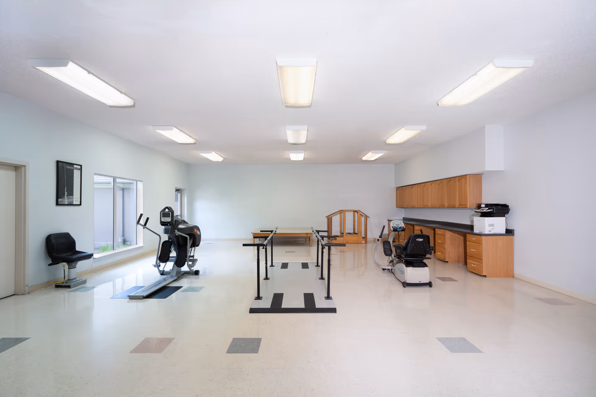 A spacious rehabilitation or physical therapy room with exercise equipment including a treadmill, stationary bike, parallel bars for walking practice, and a small set of stairs. The room has bright overhead lighting, a large window on the left wall, light-colored walls, and cabinets with a countertop along the right side.
