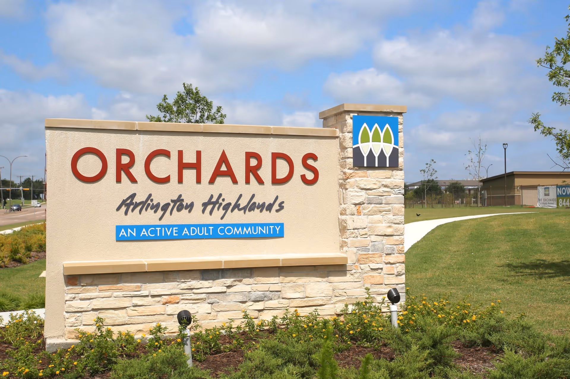 Outdoor stone and stucco sign for Orchards Arlington Highlands, an active adult community, with a landscaped area and a partly cloudy sky in the background.