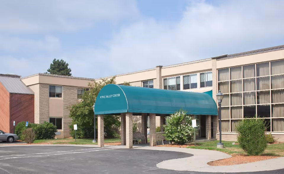 Exterior view of a two-story building with a green covered entrance labeled 'Apple Valley Center'. There are bushes and trees around the building, a parking area in front, and a clear sky above.