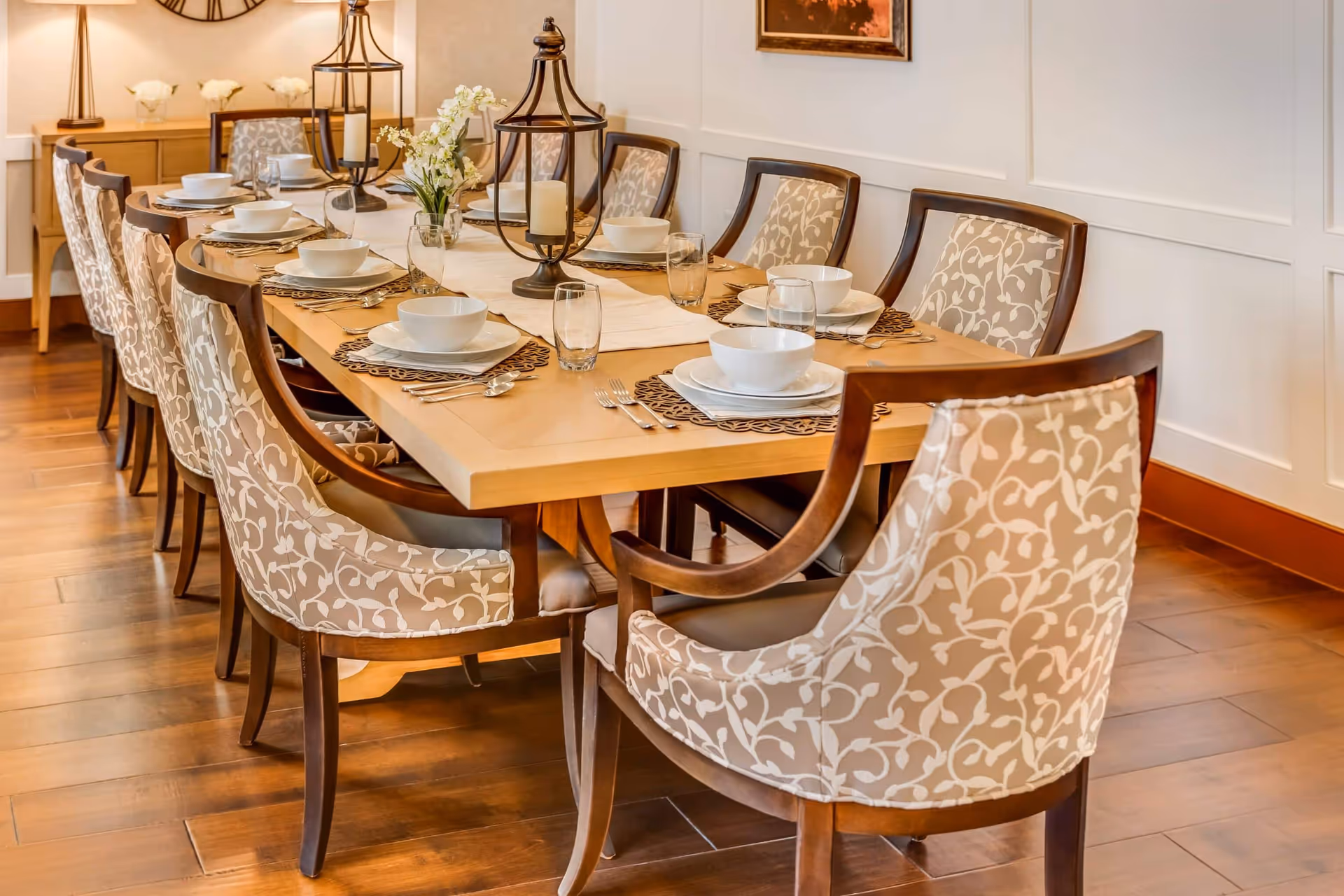 A long wooden dining table set for a meal with white bowls, plates, glasses, and silverware. The table is decorated with a white runner, two large candle lanterns, and a small floral arrangement. Surrounding the table are upholstered chairs with a beige and white floral pattern. The room has wooden flooring and white paneled walls with a framed picture hanging.