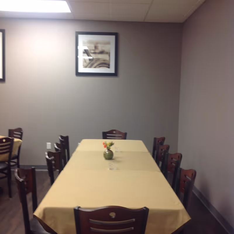 Long dining table with a beige tablecloth and a small vase centerpiece surrounded by wooden chairs in a plain room with framed art on the wall.