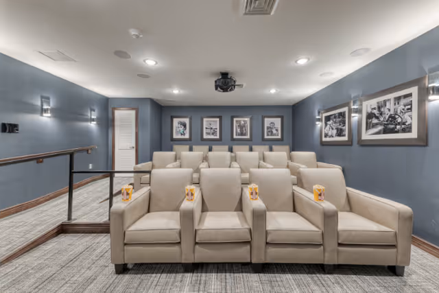 A small theater room with two rows of beige recliner chairs, each with a box of popcorn on the armrest. The walls are painted blue and decorated with black and white framed photographs. Ceiling lights and wall sconces illuminate the room, and a projector is mounted on the ceiling.