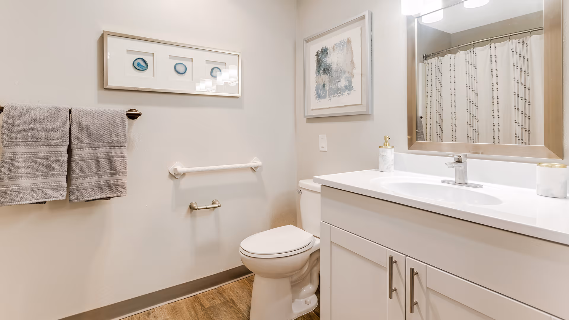 A clean and modern bathroom featuring a white toilet, a white vanity with a sink and silver faucet, a large mirror above the sink, two gray towels hanging on a towel rack, a shower curtain with a subtle pattern, and framed artwork on the walls. The floor has a wood-like finish.