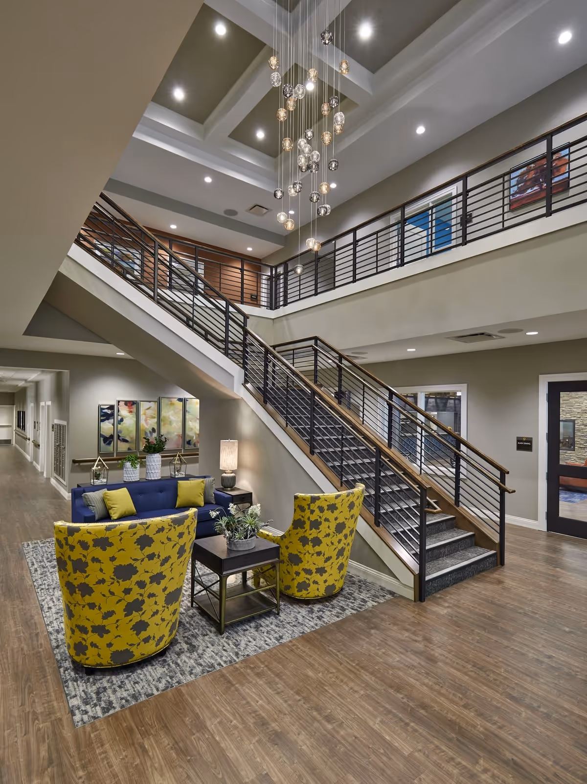 Interior view of a senior living facility lobby area with a modern staircase leading to an upper floor. The seating area includes a blue sofa with green cushions, two yellow floral-patterned armchairs, a small table with a plant, and a lamp. The ceiling features a decorative hanging light fixture with multiple glass orbs. The walls are adorned with abstract artwork, and the floor is wood with a patterned rug under the seating area.