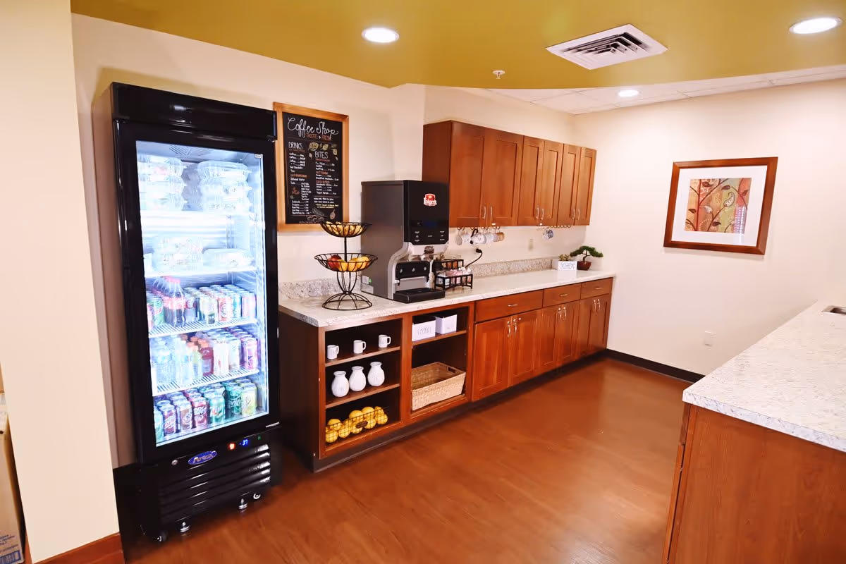 Interior view of a coffee shop area in The Cortland Wyoming facility featuring a glass-door refrigerator stocked with drinks and food items, a coffee machine on a countertop with wooden cabinets above and below, a fruit basket, mugs hanging under the cabinets, and a framed artwork on the wall.