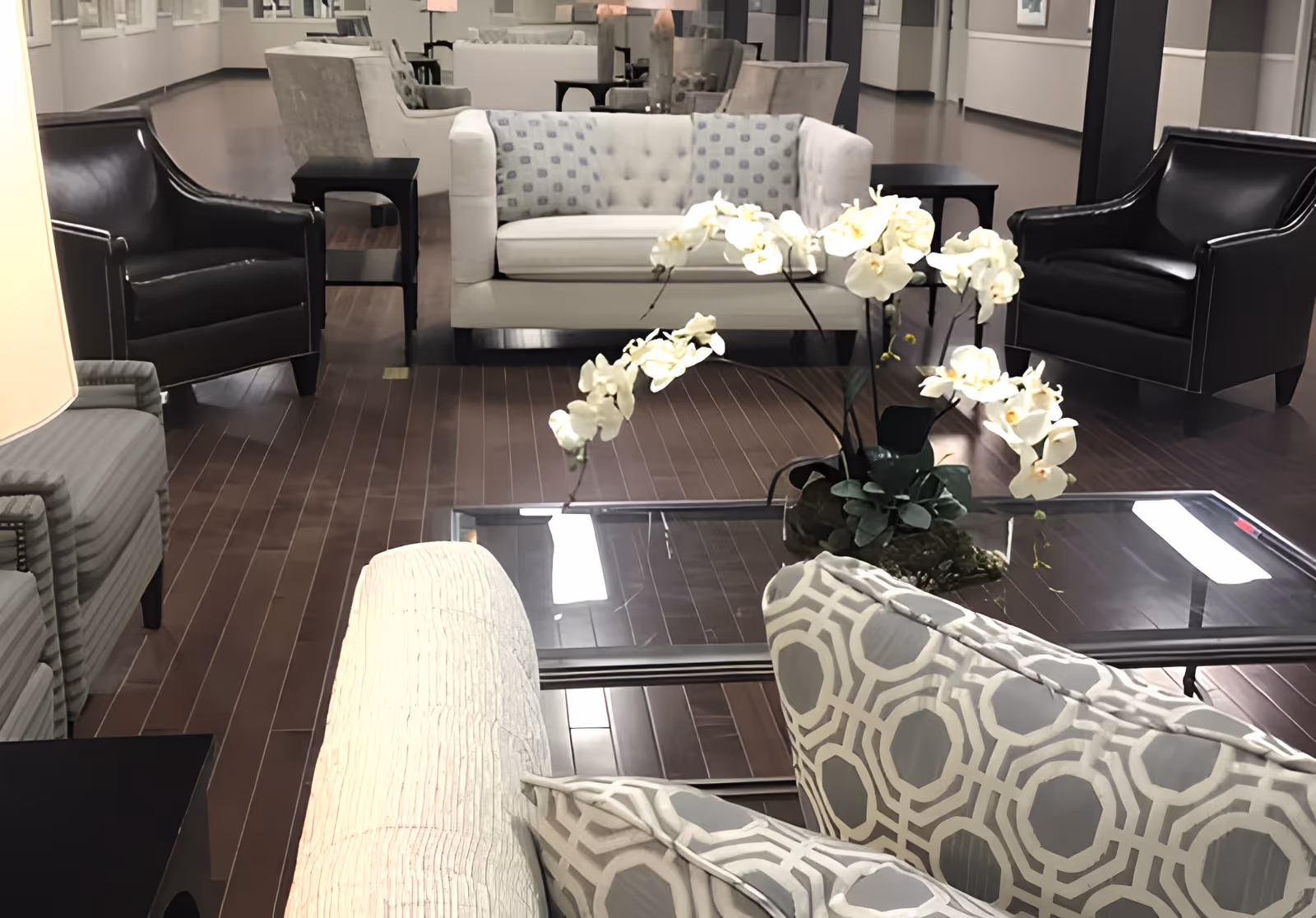 A furnished lounge with sofas, armchairs, a glass coffee table and a white orchid centerpiece on dark wood flooring.