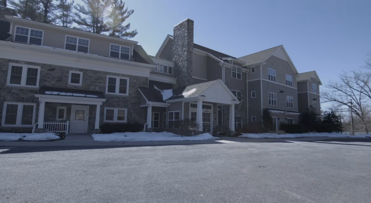 Exterior view of a senior living facility building with stone and siding facade, multiple windows, a covered entrance, and some snow on the ground under a clear blue sky.