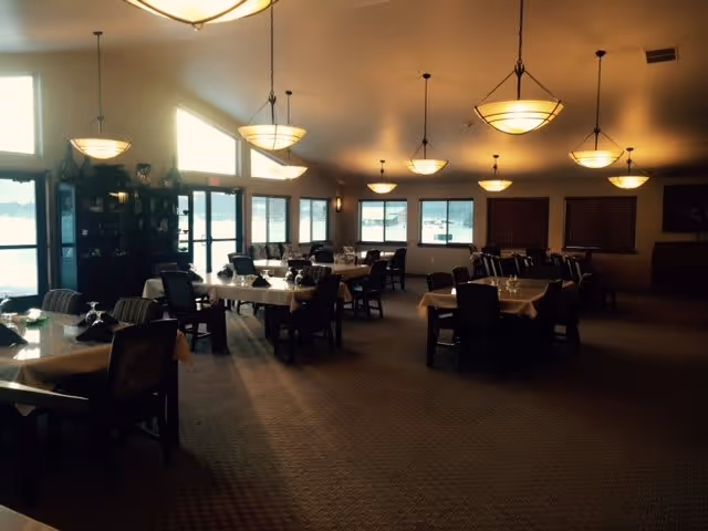 A spacious dining room with multiple tables covered with white tablecloths and set with glassware. The room has large windows allowing natural light to enter, and several ceiling lights provide additional illumination. Chairs are arranged around the tables, and there is a cabinet with plants and decorative items near the windows.