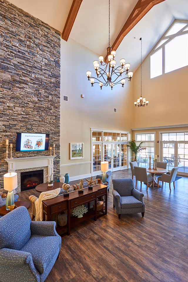 Spacious senior living facility common area with high vaulted ceilings, large windows, and wooden beams. The room features a stone fireplace with a mounted TV above it, comfortable blue armchairs, a wooden console table with decorative items, round dining tables with chairs, and elegant chandeliers hanging from the ceiling. The floor is wood, and there are glass doors leading to an outdoor area.