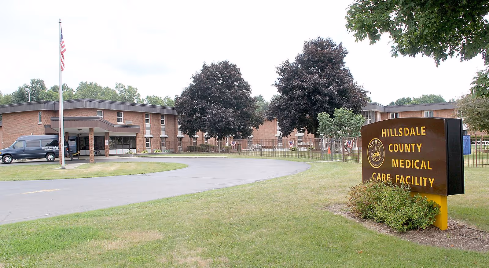 Exterior view of Hillsdale County Medical Care Facility showing a brick building with multiple windows, a driveway with a van parked near the entrance, an American flag on a flagpole, and a large sign with the facility's name surrounded by grass and trees.