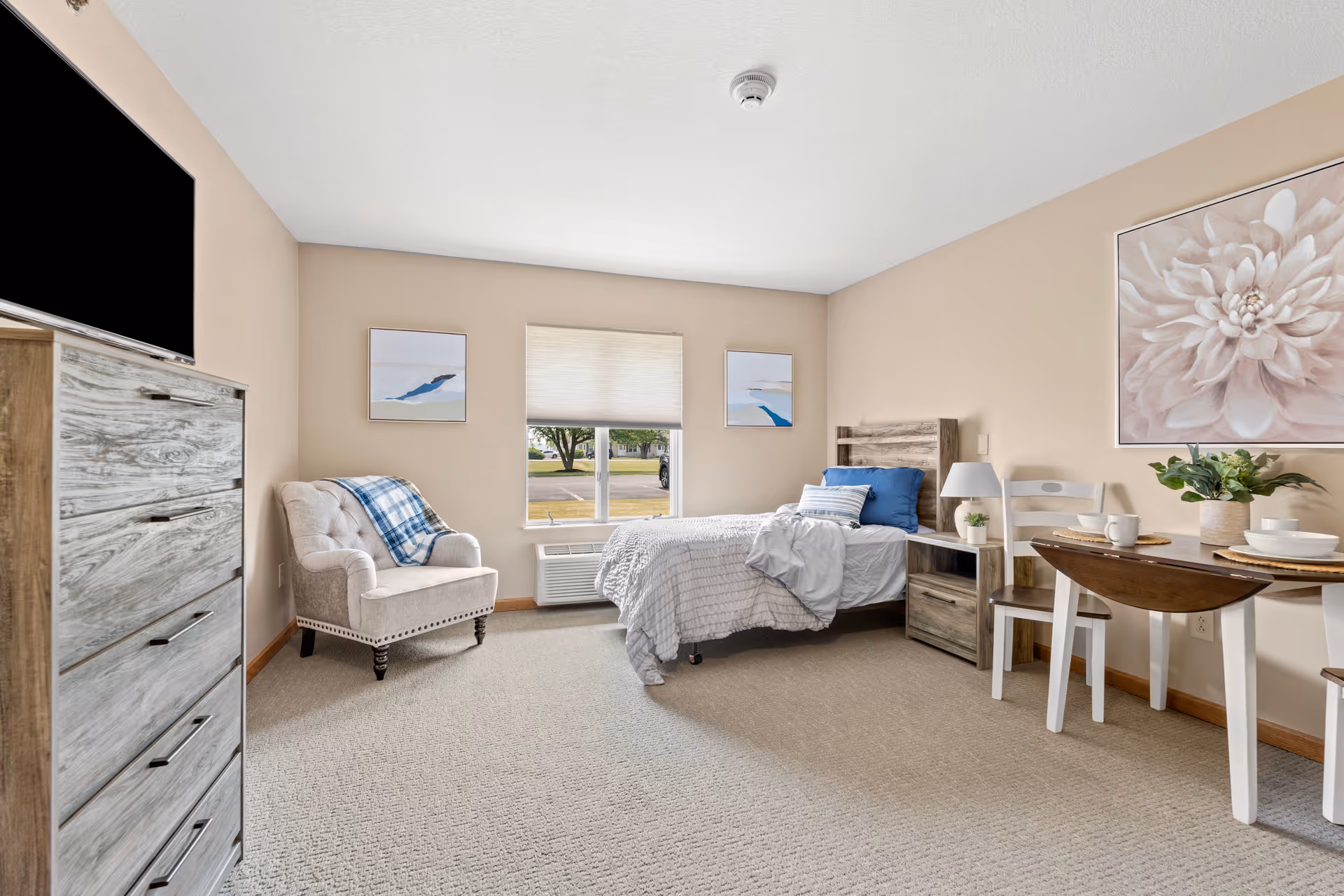 A bright and cozy bedroom in a senior living facility with a single bed dressed in white and blue bedding, a wooden nightstand with a lamp, a beige armchair with a blue plaid throw, a wooden dresser with a flat-screen TV on top, a small dining table set with plates and cups, and wall art including a large flower painting and two smaller abstract paintings. A window with a view of trees and a parking lot lets in natural light.