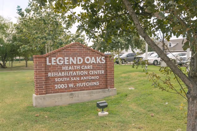 A brick sign for Legend Oaks Health Care Rehabilitation Center South San Antonio at 2003 W. Hutchins, situated on a grassy area with trees and parked cars in the background.