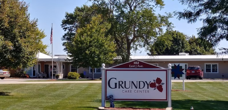 Exterior view of Grundy Care Center building with a large sign in the foreground displaying the facility name and address. The building is single-story with several windows, surrounded by green grass, trees, and a clear blue sky. An American flag is visible near the entrance.