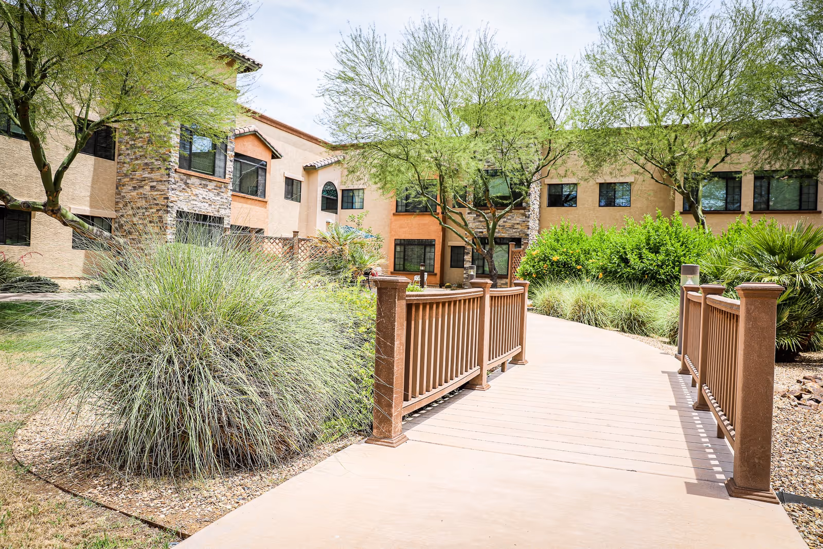 A sunny outdoor pathway with a wooden railing bridge leading to a two-story building with beige and orange walls, surrounded by green trees and bushes.