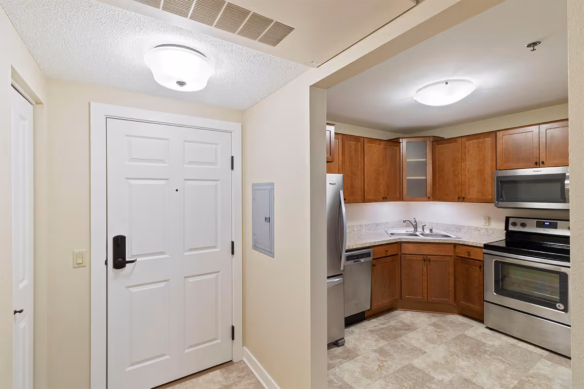 Interior view of a senior housing unit showing the entrance door on the left and a kitchen area on the right with wooden cabinets, a stainless steel refrigerator, dishwasher, stove, microwave, and a corner sink.