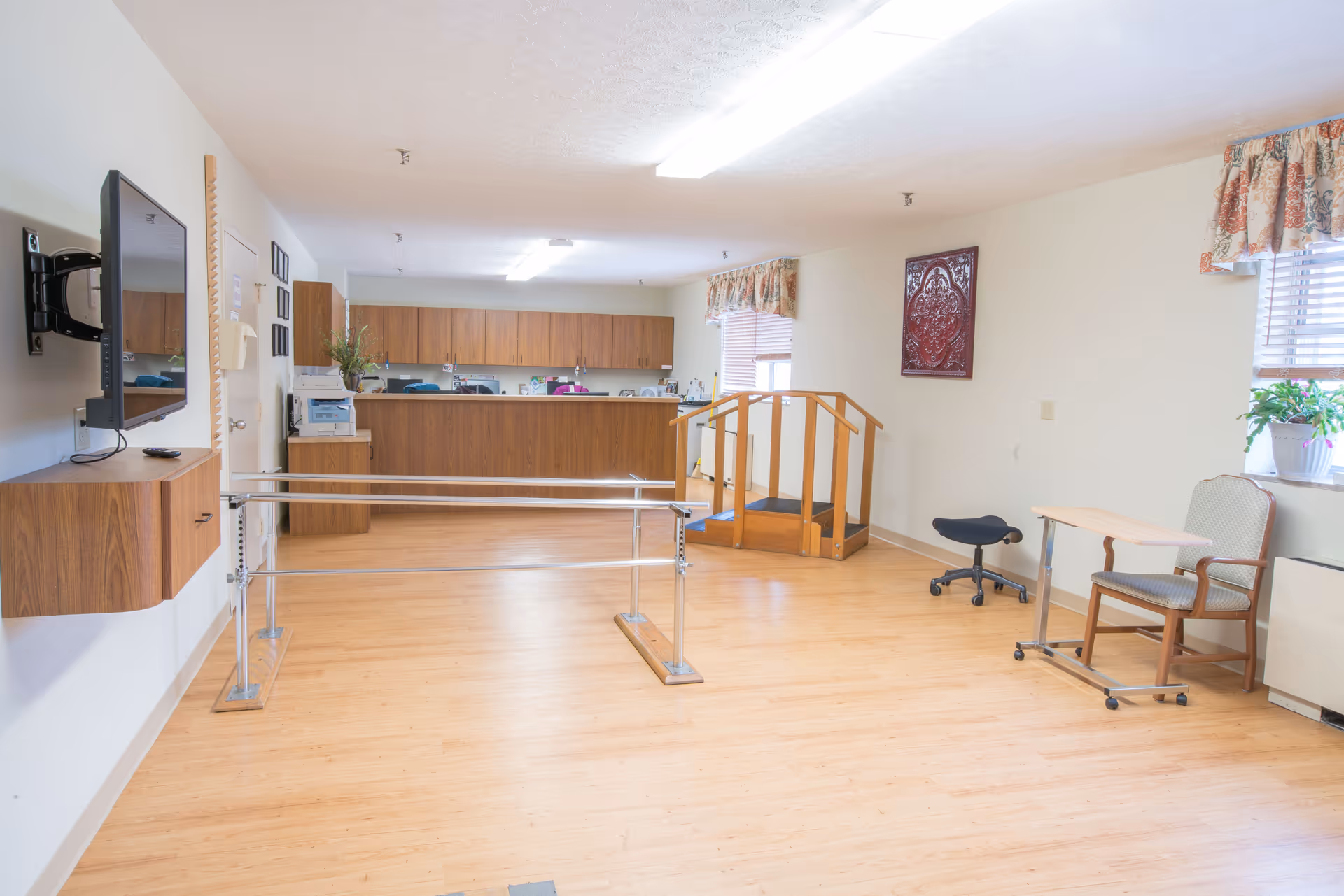 A spacious room with wooden flooring featuring parallel bars for walking exercises, a small wooden ramp with handrails, a chair with a rolling tray table, a stool, and a wall-mounted TV. The back of the room has a counter with cabinets above and below, and windows with floral valances letting in natural light.