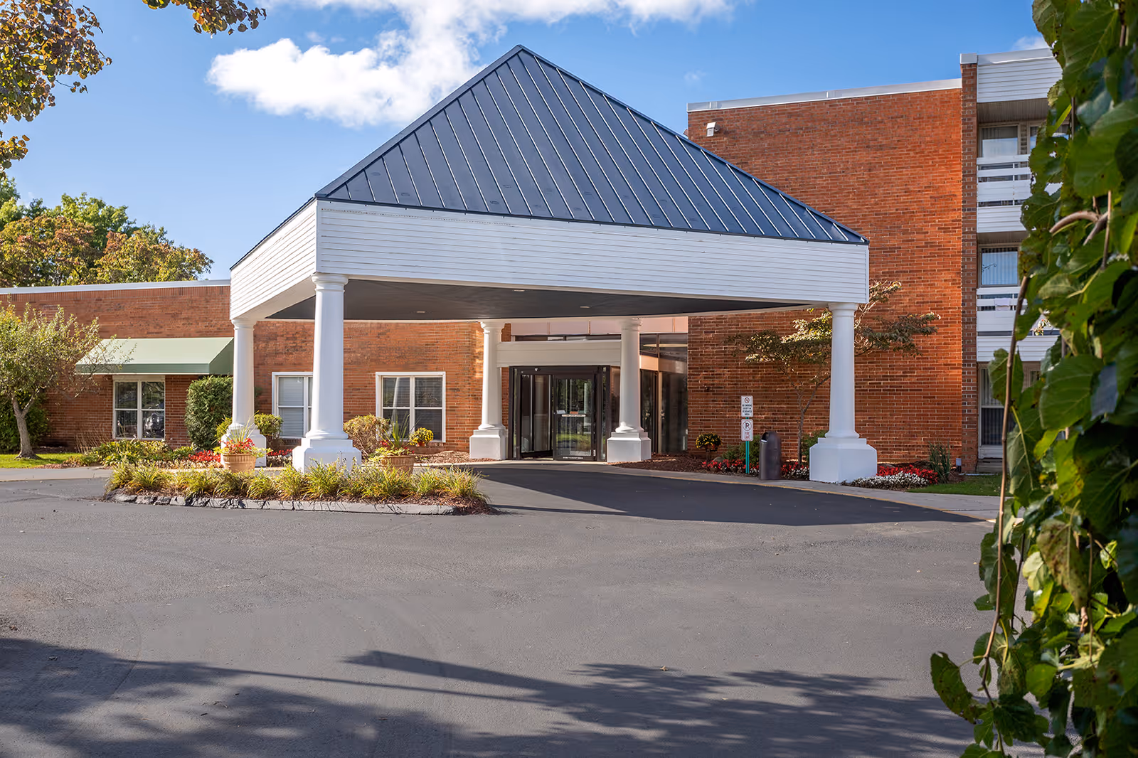Entrance of a senior living facility named Park At Franklin with a covered drop-off area supported by white columns, surrounded by landscaped plants and a brick building under a blue sky with some clouds.