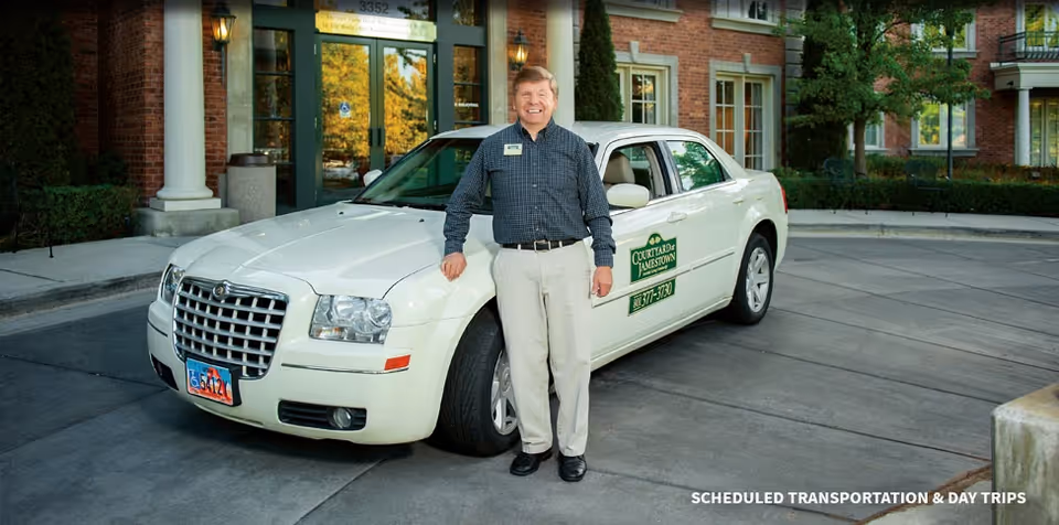 A man standing and smiling next to a white car with a Courtyard at Jamestown logo on the door, parked in front of a brick building entrance with large windows and columns.