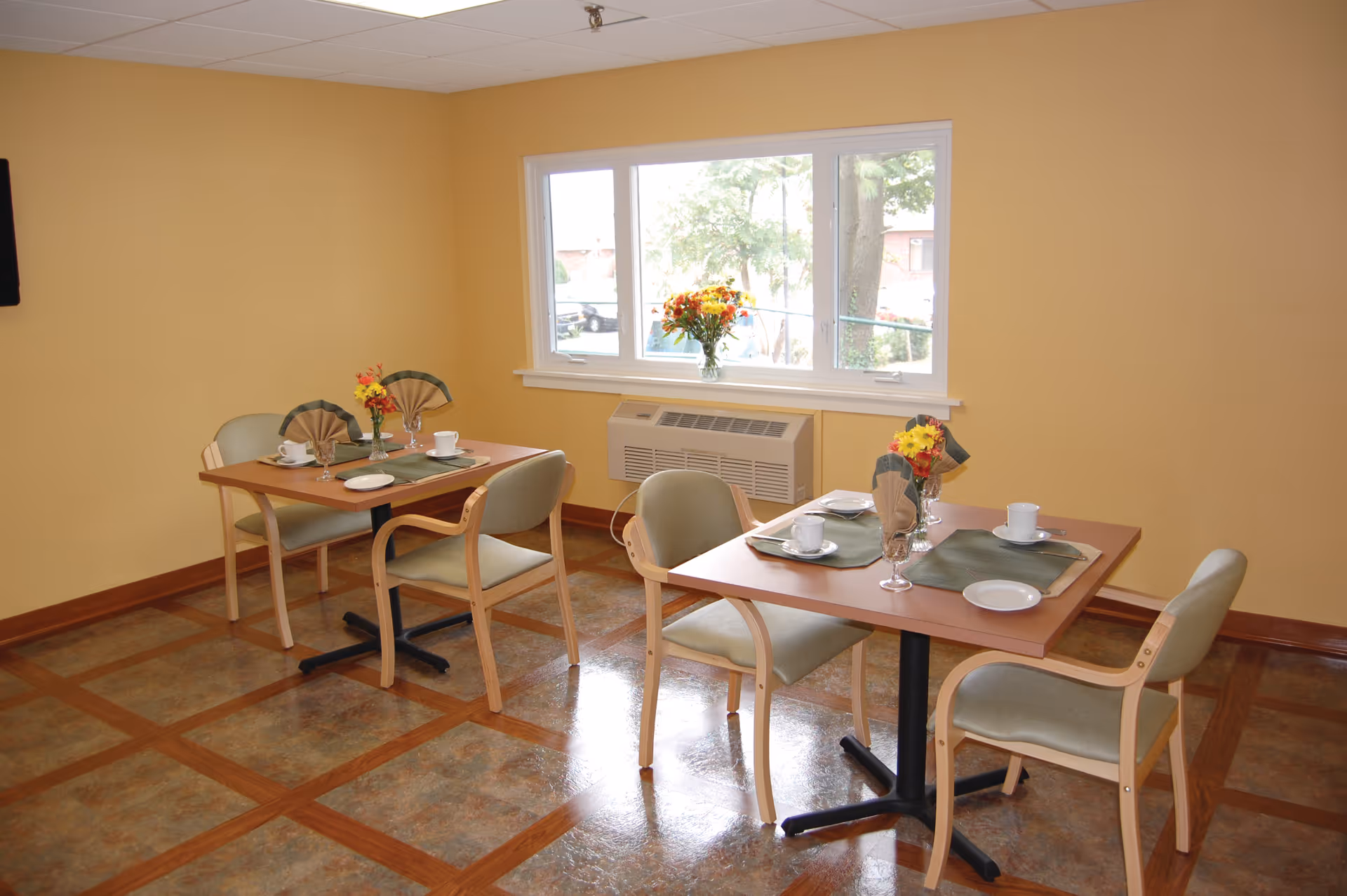 A small dining area with two square wooden tables, each set with placemats, napkins, cups, and plates. Four cushioned chairs with wooden frames surround the tables. A window with a view of trees and a building outside is centered on the yellow wall, with a vase of flowers on the windowsill. The floor has a patterned tile design with wooden borders.