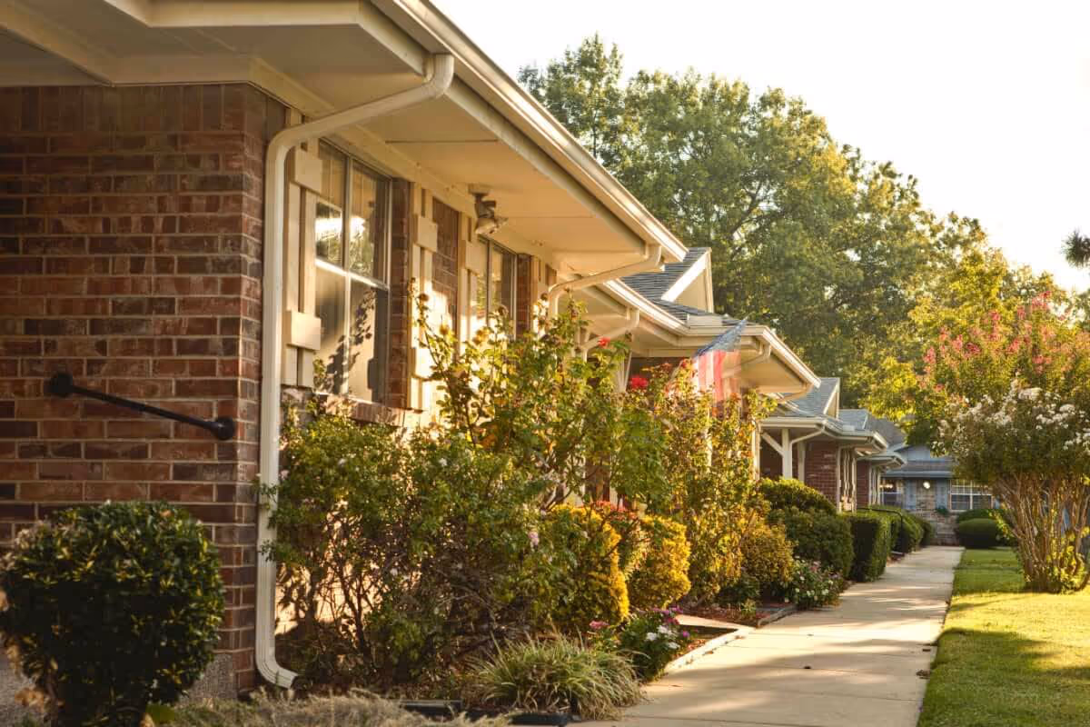 A sunny walkway lined with bushes and flowering plants alongside a row of brick and siding residential buildings with windows and porches, with trees in the background.