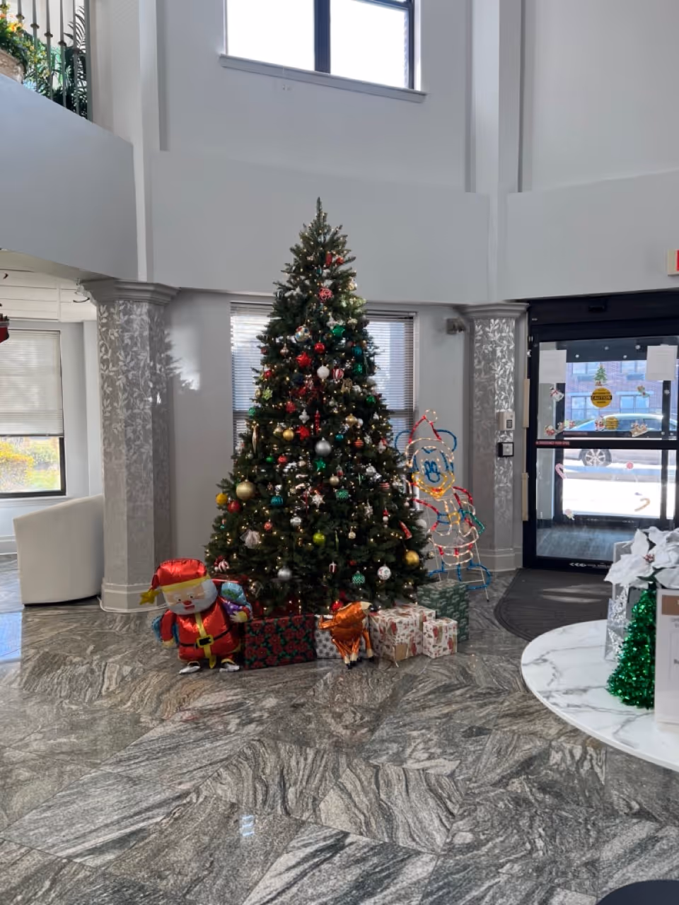 A decorated Christmas tree with ornaments and lights stands in the center of a spacious lobby area with marble flooring. Wrapped presents and holiday decorations, including an inflatable Santa and a light-up figure, are placed around the tree. Large windows and a glass door letting in natural light are visible in the background.