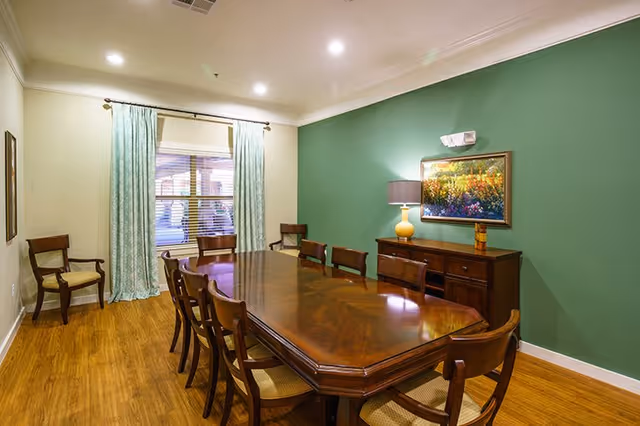 A dining room with a long wooden table surrounded by eight wooden chairs. The room has wood flooring, a green accent wall with a painting and a lamp on a sideboard, and a window with light blue curtains and blinds.