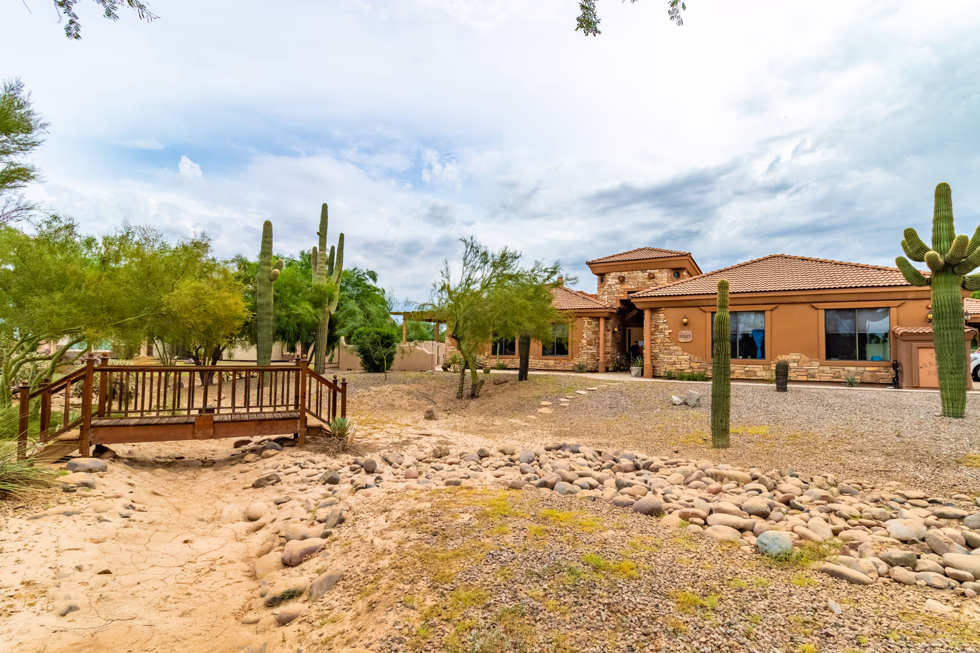 Exterior view of Crismon Peaks Assisted Living facility showing a desert landscape with cacti, trees, rocks, and a small wooden bridge over a dry creek bed. The building has a southwestern architectural style with stone and stucco walls and a tiled roof under a partly cloudy sky.