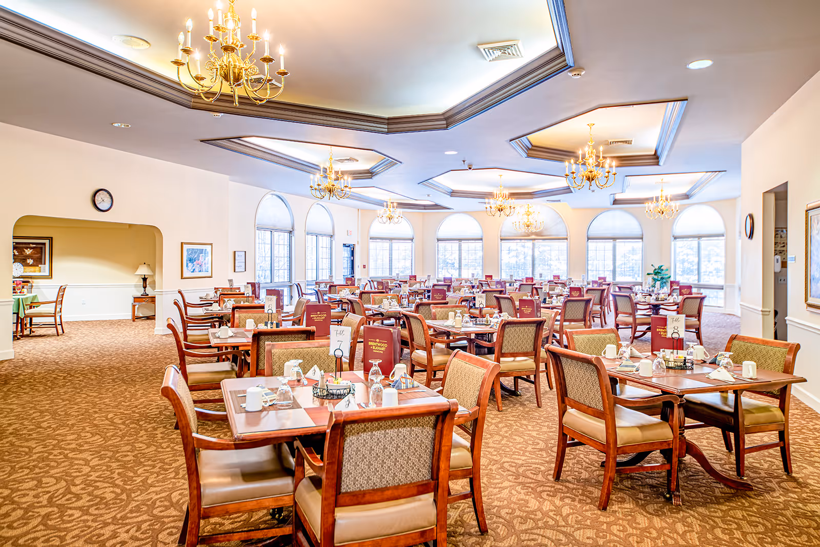 A spacious and well-lit dining room in an assisted living facility with multiple wooden tables and chairs arranged neatly. Each table is set with placemats, glasses, cups, and menus labeled Brentwood at Elkhart. Large arched windows allow natural light to fill the room, and elegant chandeliers hang from the ceiling.