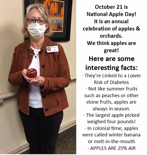 A woman wearing glasses, a face mask, and a brown jacket stands indoors holding a red apple in her hands. Behind her is a framed picture on the wall. To the right of the image is text about National Apple Day on October 21, listing interesting facts about apples.