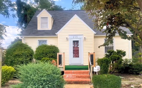 Front exterior view of a small yellow house with a dark gray roof, a white door with the number 215 above it, brick steps leading up to the entrance, and green bushes and trees surrounding the house.