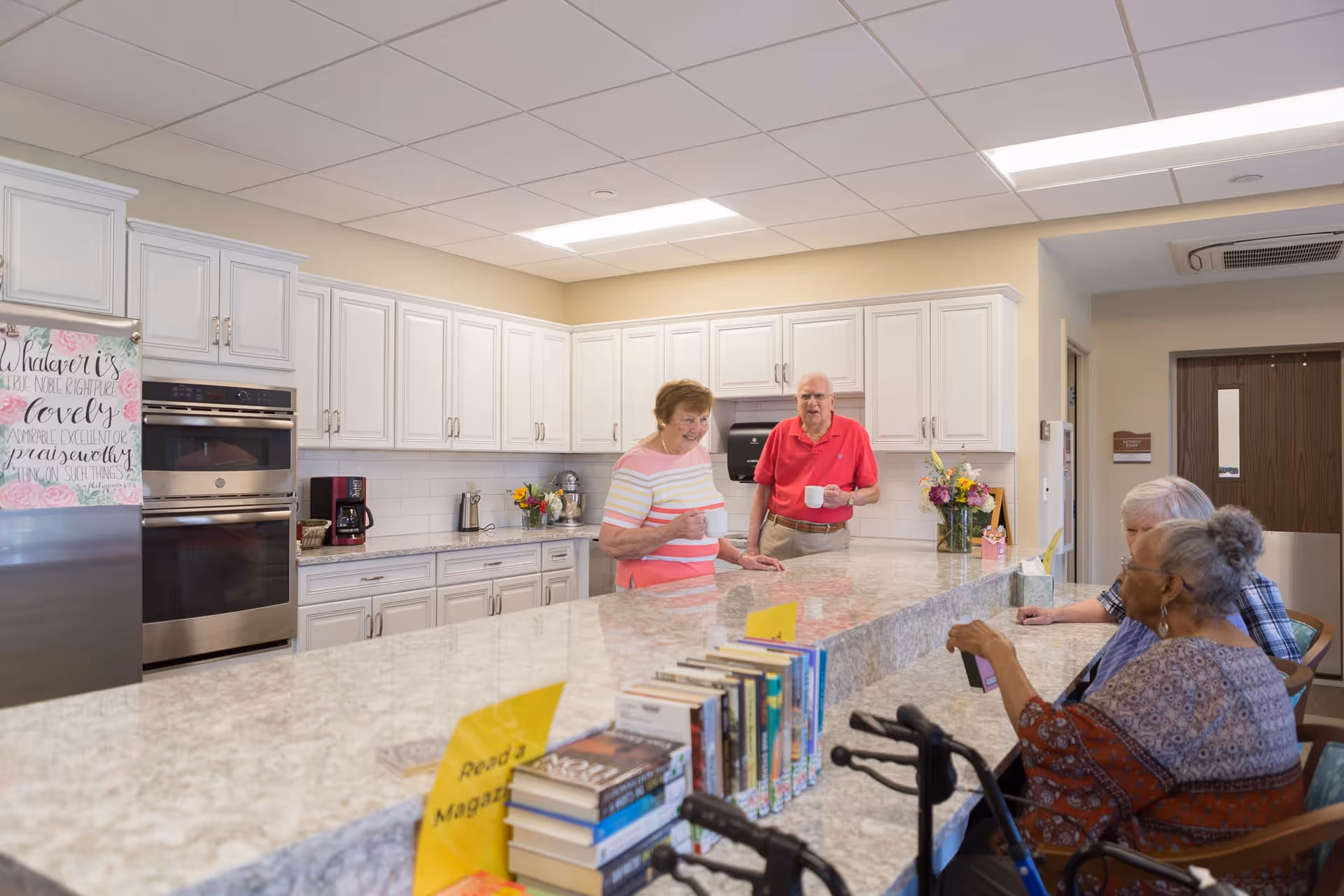 Several senior residents chatting around a large marble island in a bright communal kitchen with white cabinets and appliances.