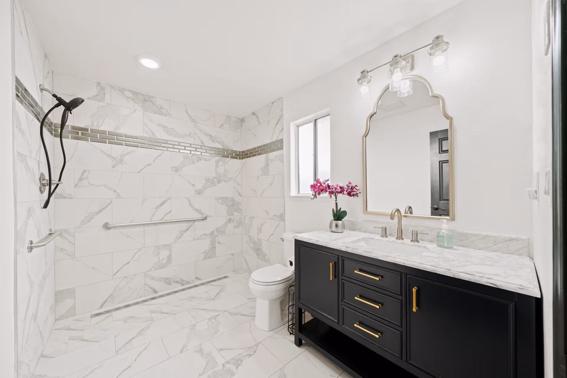 A modern bathroom with white marble tiles on the floor and walls, featuring a walk-in shower with a handheld showerhead and grab bars. There is a white toilet next to a black vanity with a marble countertop, gold handles, a decorative pink orchid, a soap dispenser, and a large ornate mirror above it. The bathroom is well-lit with ceiling and vanity lights.