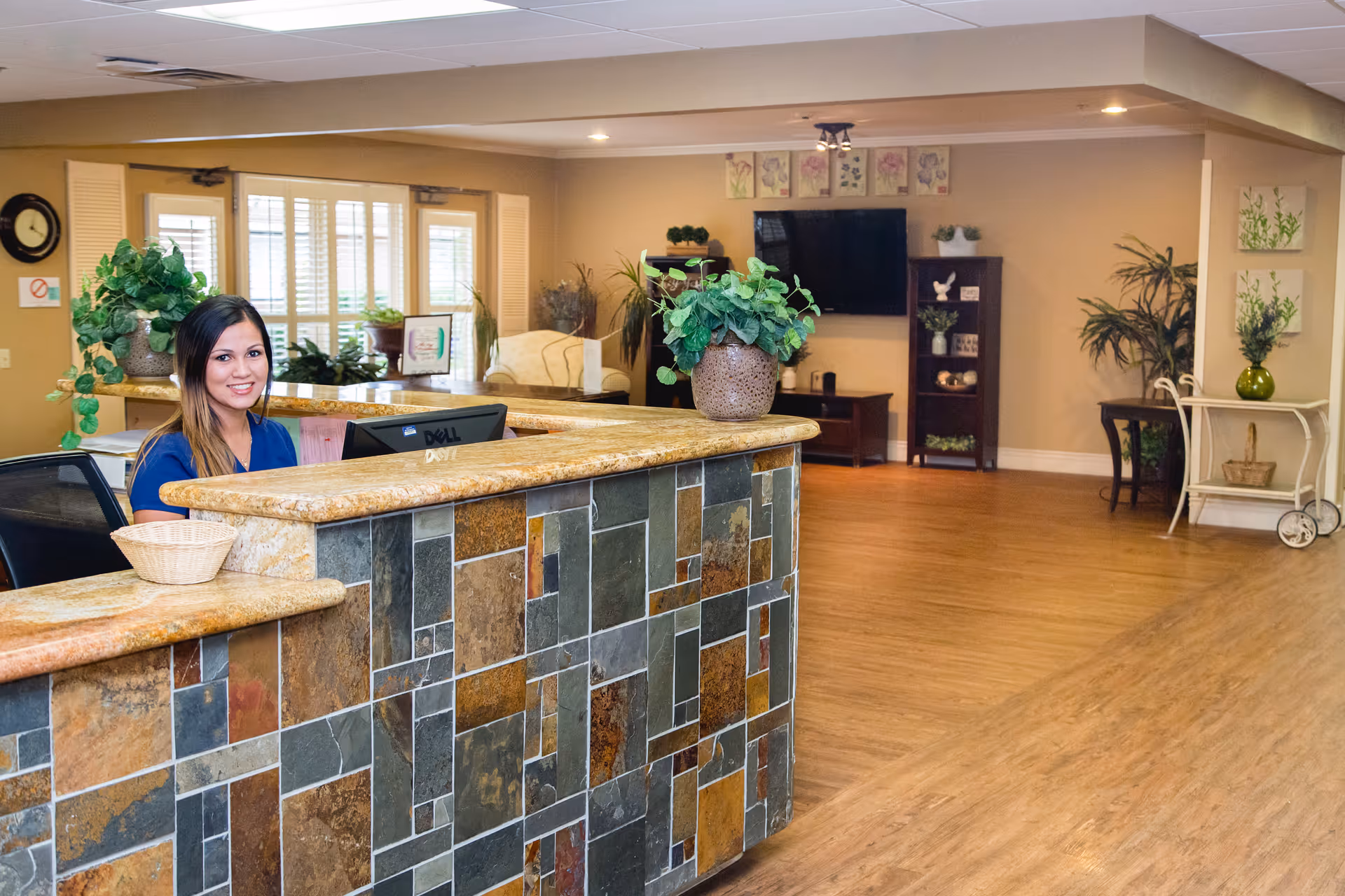 Reception area of Palm Valley Post Acute facility with a smiling receptionist seated behind a stone-tiled counter. The room has wooden flooring, potted plants on the counter and around the room, a wall-mounted TV, shelves with decorative items, and large windows with blinds letting in natural light.