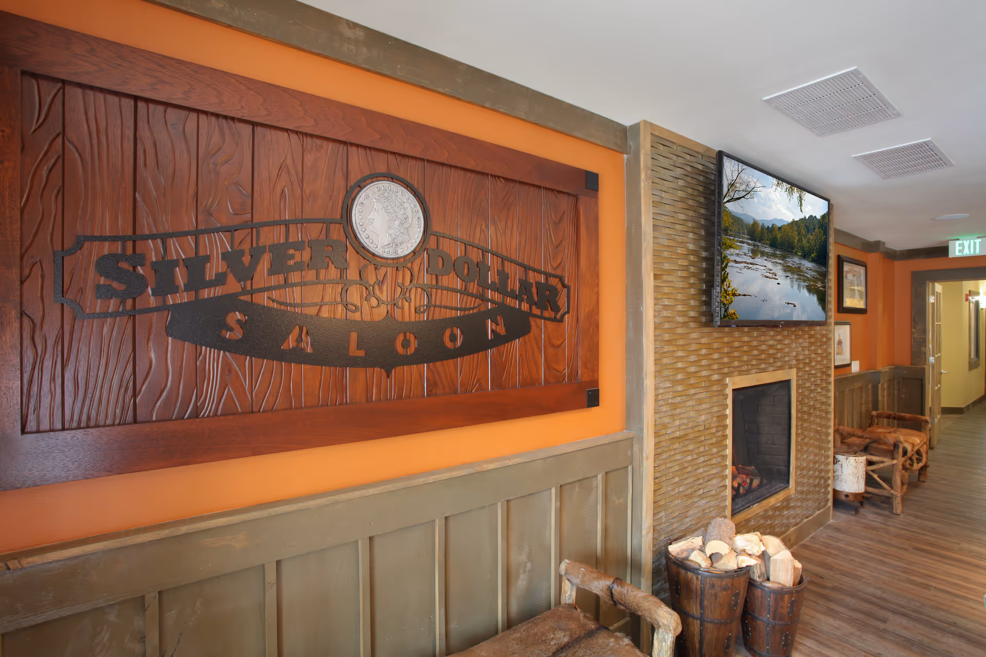 Interior hallway of a senior living facility with rustic decor including a wooden sign reading 'Silver Dollar Saloon', a fireplace with stacked firewood, a mounted flat-screen TV displaying a nature scene, wooden chairs, and framed pictures on the walls.