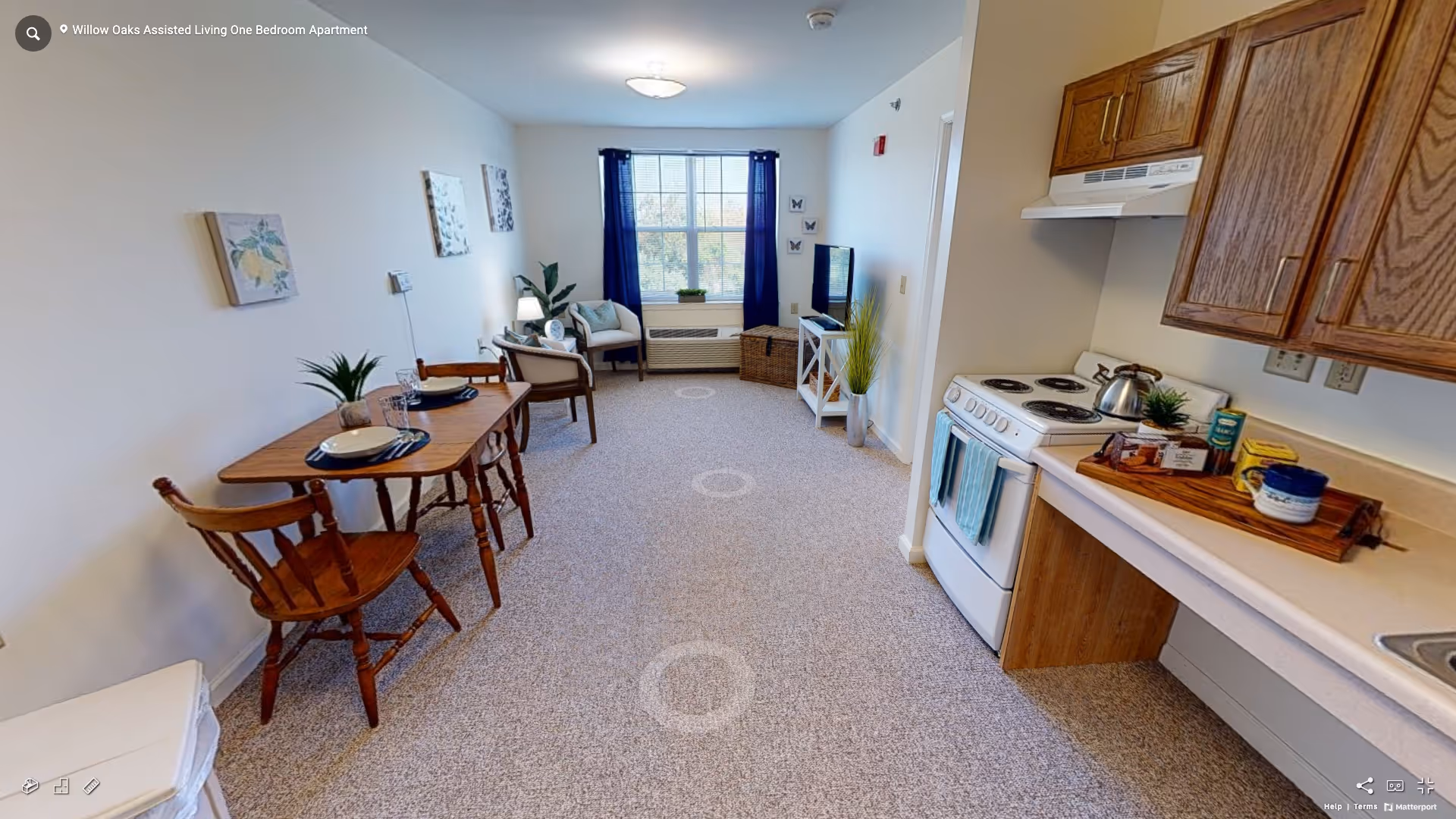 A bright and cozy assisted living apartment featuring a small dining table with two wooden chairs on the left, a compact kitchen area with stove and cabinets on the right, and a living area with a chair, small table, and TV near a window with blue curtains at the far end.