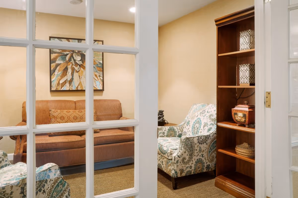 View through glass‑paneled doors into a cozy sitting room with a sofa, patterned armchairs, wall art, and a wooden bookshelf.
