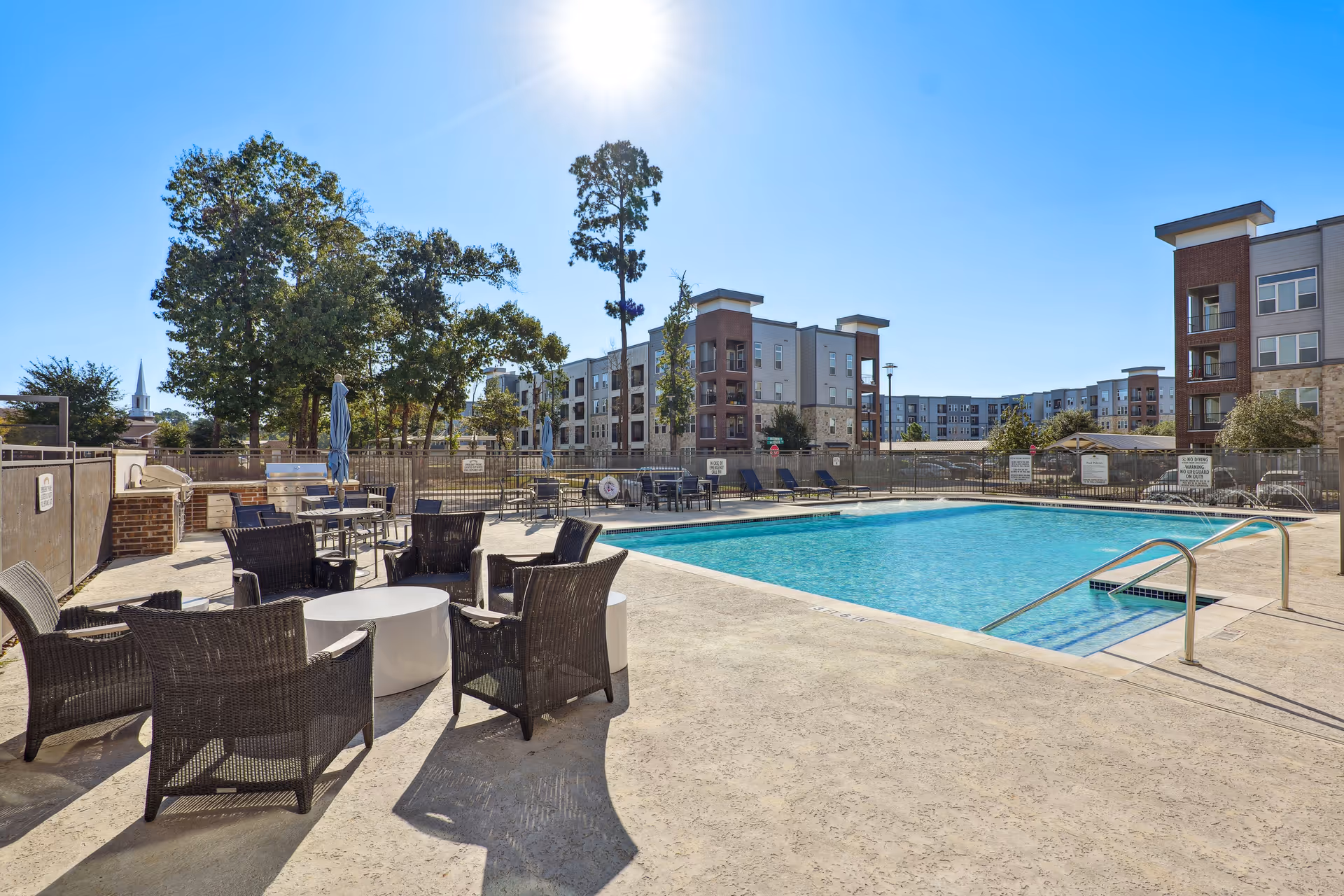Outdoor swimming pool area with clear blue water, surrounded by patio chairs and tables. Several multi-story residential buildings are visible in the background under a bright blue sky with the sun shining. Trees and umbrellas are also present around the pool area.